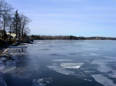 [A view from the dock such that the shore in on the left and in the far distance. Deep blue water with floating sections of thin ice comprises most of the image. Blue sky fills most of the remainder of the image.]